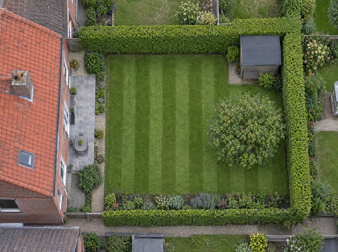 Vue du ciel d’un jardin derrière une maison de briques : pelouse aux bandes de tonte claires et foncées, haies taillées, abri de jardin et pommier